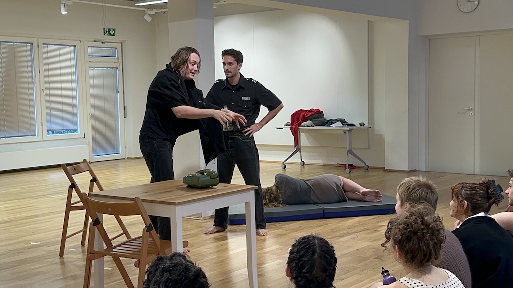 Two actors stand next to one another in mid-performance of 'Faith Hope Charity' while a third actor lays on a mattress off to the right. Students are seated in the audience watching the performance.